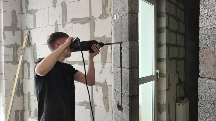 A man drills a hole in a wall, preparing to install electrical boxes. The scene takes place in the living room during a home renovation. The work is being done during the day.