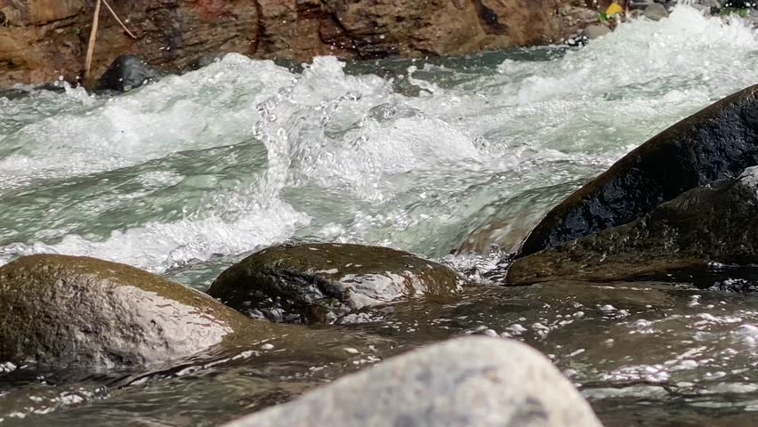 Close-up of powerful river rapids flowing over rocks in a mountain stream. A high-angle, close-up shot of crystal clear water rushing over large stones in a fast-flowing river. The image captures the dynamic movement of white water foam and splashes against smooth river rocks, creating a refreshing and natural atmosphere. Perfect for themes related to nature, environment, clean water resources, and outdoor adventure.