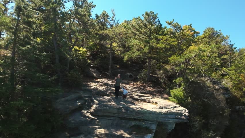 White Woman Hiking On Cliff Ledge Stopping To Admire Forest Panorama, Rocky Shelf Underfoot, Pine Trees Framing Bright Blue Sky, Peaceful Solitude And Soft Daylight Mood