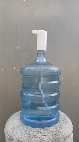 Close-up of a person filling a glass with water from a large gallon bottle using an automatic electric pump dispenser in a room.