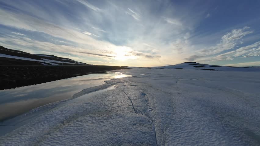 An FPV drone glides low along the edge of the frozen lake, flying into the sun as warm light spills across the ice, creating glowing reflections and a beautifully backlit winter scene.