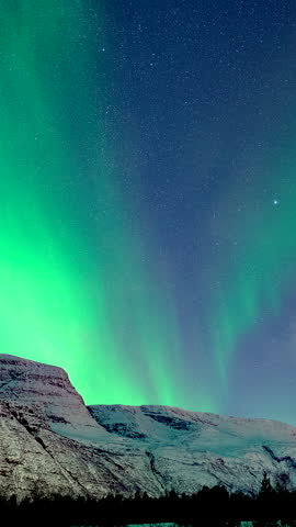 Vivid green aurora borealis dancing across starry night sky above snowy mountains in Norway, vertical timelapse