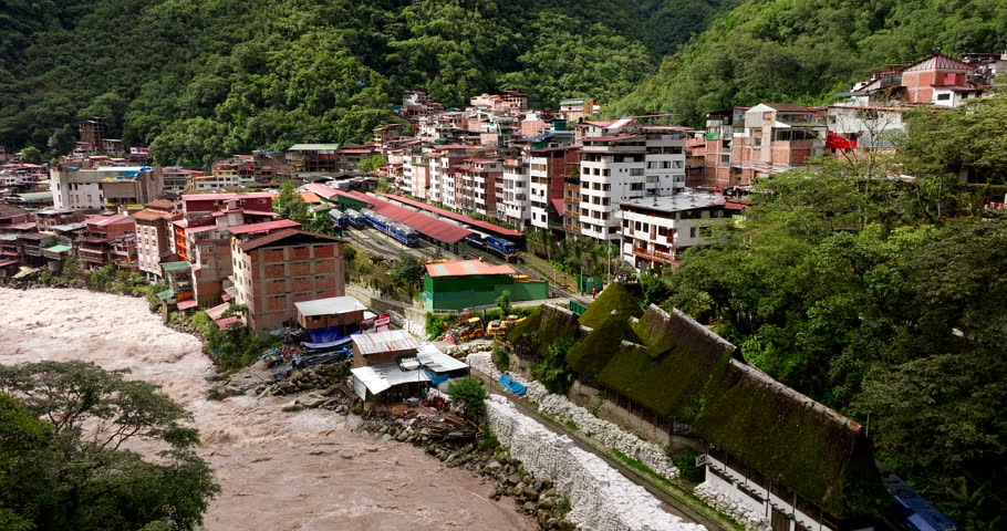 Cinematic drone glide over Aguas Calientes, Peru, as a swollen rapid river surges through the valley below Machu Picchu, rushing past dense hillside buildings under lush jungle slopes