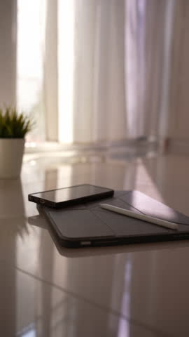 Vertical shot of smartphone on digital tablet with stylus and ceramic plant pot, sunlight through blowing curtains by glass door, cozy morning workspace, 4K.