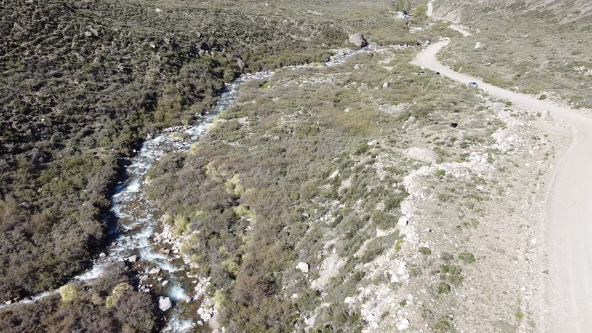 Drone view of a river flow through a rugged landscape near a dirt road in Cajon de Arenales, Mendoza, Argentina. Great for travel, nature