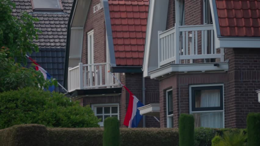 Row of brick houses in Dutch neighborhood displaying national flags at half mast. Traditional commemoration of war victims on May 4. Respectful atmosphere in typical Netherlands suburb.
