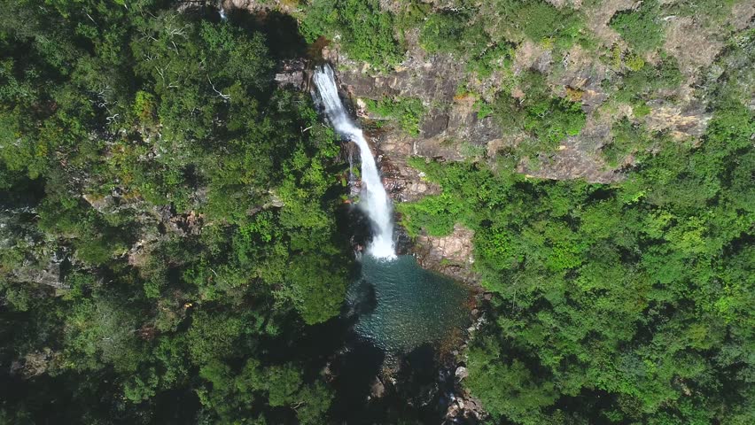 Aerial drone view of Ouzoud Falls in Morocco showing tall waterfall flowing through rocky cliffs into clear pool below, surrounded by lush green forest and dramatic natural landscape