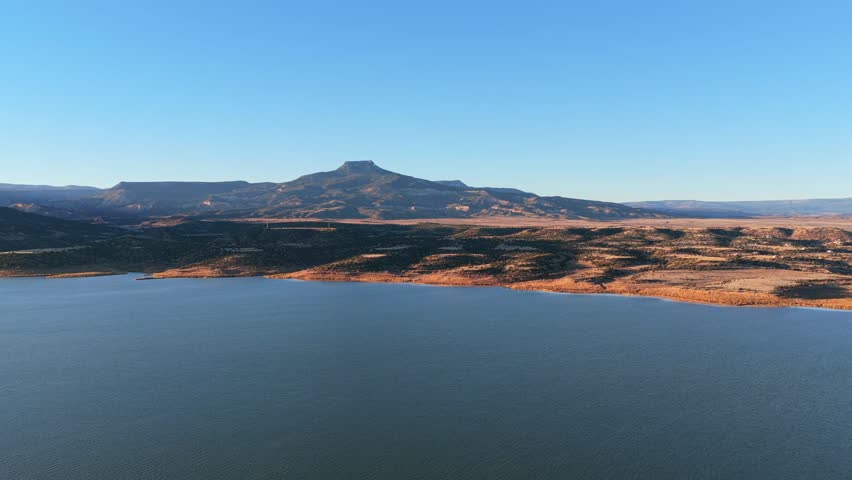 Deep blue water of Abiquiu Lake reservoir borders the arid desert brush of northern New Mexico. The volcanic basalt peak of Cerro Pedernal rises in the distance under a clear and bright blue sky.