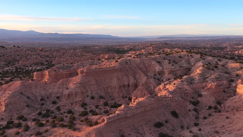 Sunlight illuminates the textured sandstone ridges and deep gullies of an ancient high desert badland. The arid landscape of New Mexico features layered earth formations and sparse green shrubs.