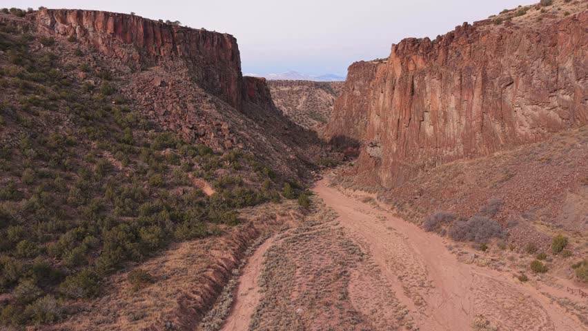 Volcanic basalt rock walls tower over the sandy wash floor within Diablo Canyon. The high desert plateau landscape of New Mexico contains arid brush and dark volcanic earth formations.