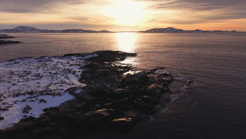 Stunning wide shot of a golden sunset reflecting on calm arctic waters. The scene features a rugged, snow-dusted rocky coastline under a dramatic purple and orange twilight sky. City Nuu, Greenland.