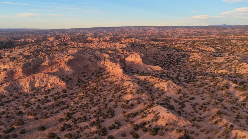 Sunlight hits the eroded ridges of a high desert badland while mountains sit on the far horizon. This wide aerial view captures the textured earth and arid beauty of the Southwest.