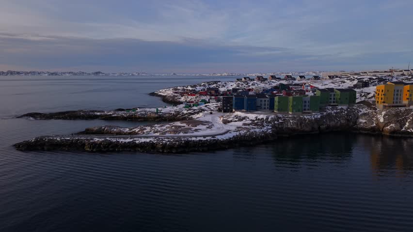 Traditional and modern colorful houses nestled on the rocky shores of Nuuk. The golden hour light illuminates the snowy landscape and the calm fjord waters.