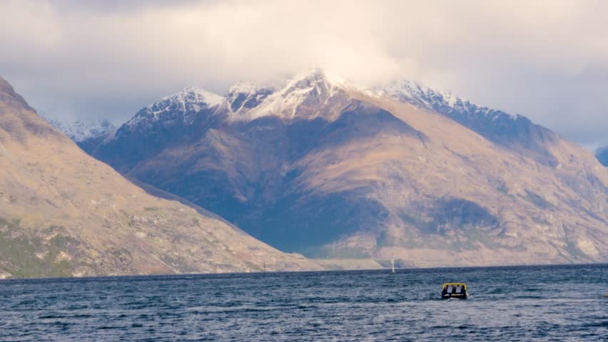 Slow motion landscape of speedboat in inlet channel bay of Lake Wakatipu surrounded by snowy ice capped mountain range summit and foggy sky over peak near Queenstown Otago New Zealand tourism nature