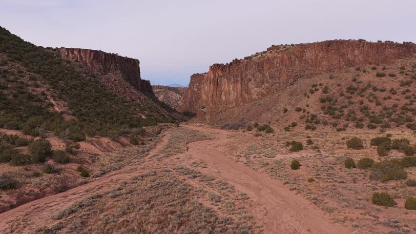 A wide aerial view shows the entrance road and parking area leading to Diablo Canyon. The high desert landscape features towering volcanic basalt rock walls and arid brush in northern New Mexico.