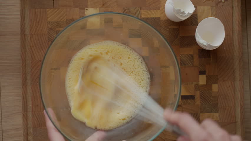 Person whisking eggs in glass bowl with metal whisk on wooden cutting board in kitchen, top view. Concept of breakfast preparation and home cooking