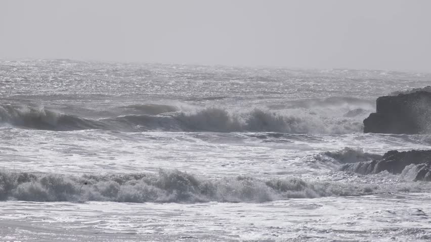 Powerful Atlantic Ocean surf breaks against dark coastal rocks, sending spray into windy air. Dramatic seascape with rough water and white foam.