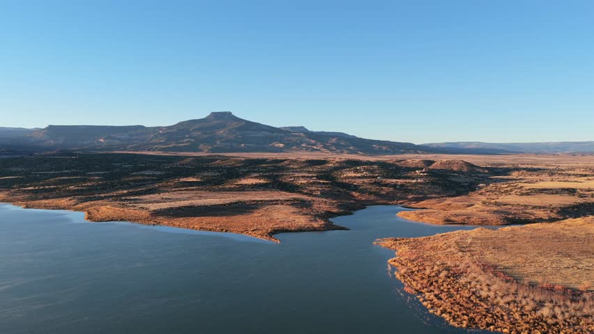 The blue water of Abiquiu Lake reservoir sits below the volcanic basalt peak of Cerro Pedernal. The high desert plateau landscape features sunlit soil and arid shorelines at the base of the mountain.