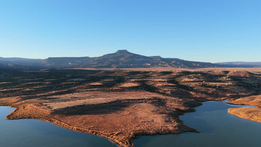 Morning sunlight hits the volcanic basalt peak of Cerro Pedernal above Abiquiu Lake. The high desert plateau landscape features arid soil and a rippled blue reservoir in northern New Mexico.