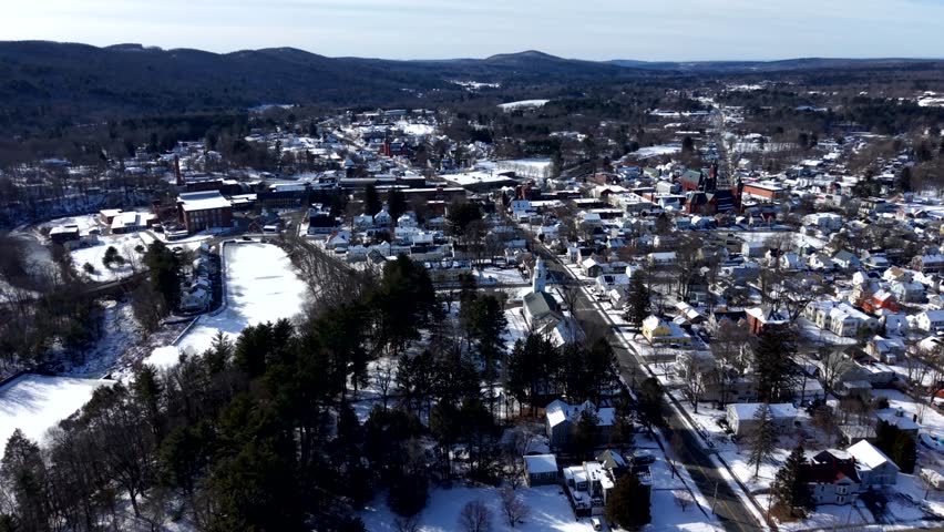 Aerial view of downtown Ware, Massachusetts in winter, showing snow covered houses, streets, trees and surrounding hills under clear daylight with long shadows and town layout. Wide shot.