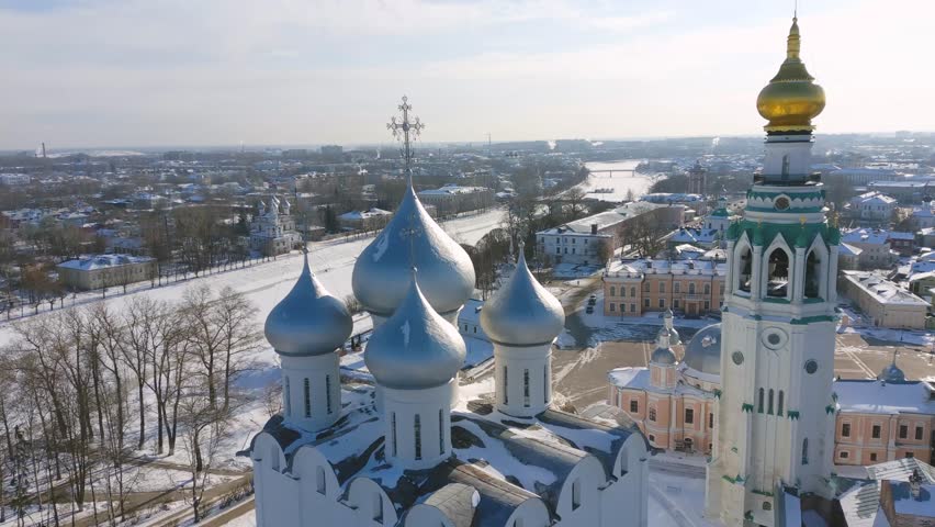 Close aerial over Vologda Kremlin Cathedral with green spire and golden dome, blue onion cupolas, white walls, overlooking frozen river and snowy Soviet cityscape, Russia.
