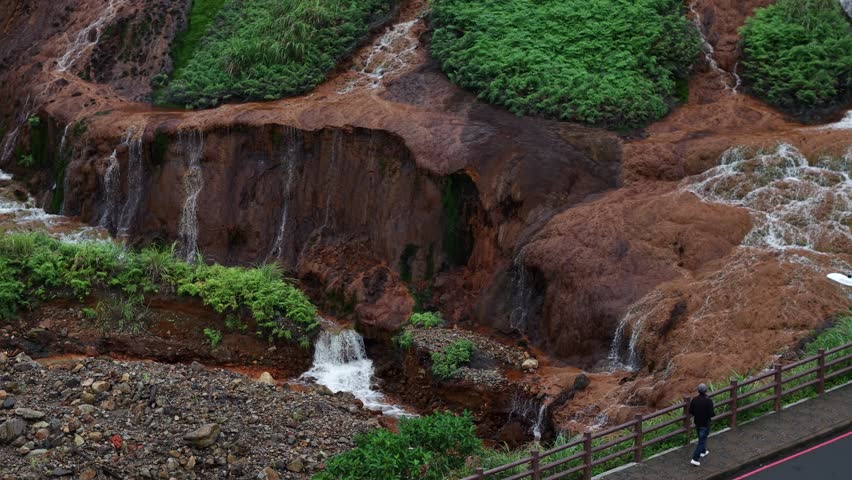 A cinematic crane shot reveals the unique Golden Waterfall in Jinguashi, Taiwan, showcasing its vibrant mineral-stained rock and cascading streams under an overcast sky.