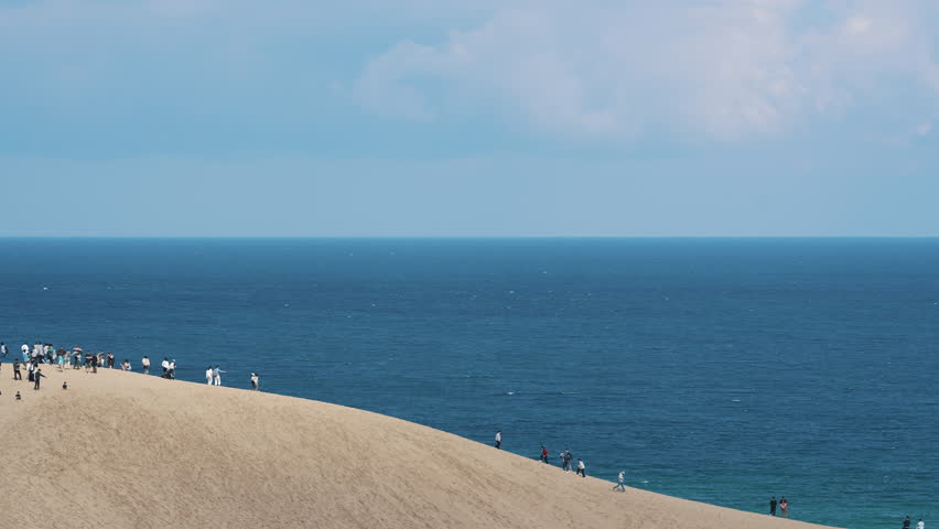 Crowds of tourists climbing the massive Tottori Sand Dunes overlooking the blue sea.