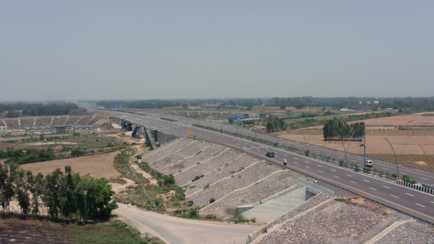 Rare overhead captures car driving on expansive multi-lane Greenfield Corridor highway, crossing modern flyover bridge amid agricultural fields and sparse vegetation under hazy conditions.