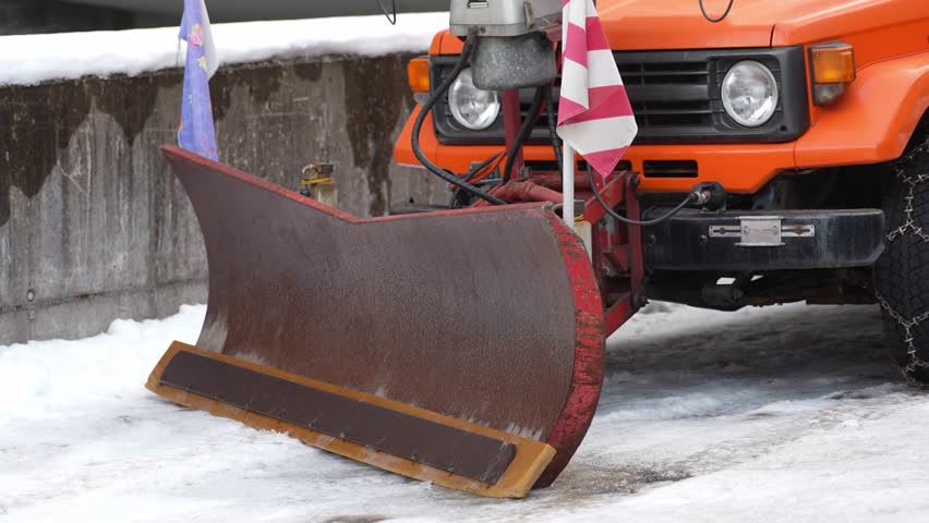 Rusty snowplow blade attached to a vintage orange off road truck parked on a snowy street