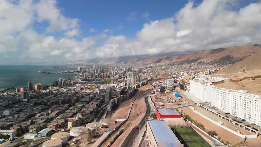Aerial pull-back drone shot of Antofagasta city, Chile, revealing urban landscape, desert hills, and the Pacific Ocean under a dynamic sky. Cinematic aerial footage showing residential neighborhoods,