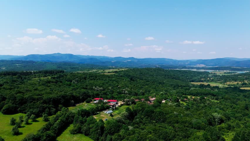 Wide aerial pan revealing Balkan hills, greenery, houses, villages, a lake, and distant hazy mountains under blue skies.