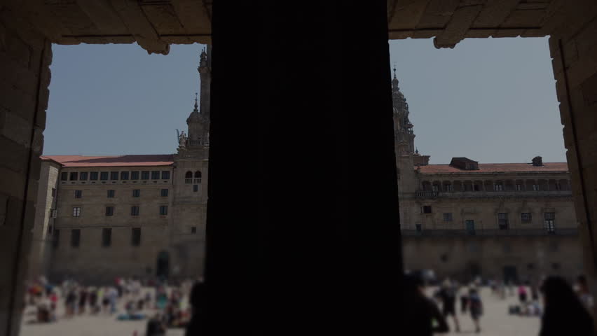 Establishing shot of Obradoiro Square framed by cathedral arches, with pilgrims softly blurred in the background.