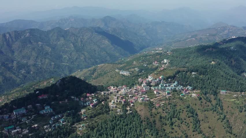High drone shot captures Mashobra village in Himachal Pradesh with cluster of colorful rooftop houses nestled amid pine-covered hills and distant hazy mountain ranges.