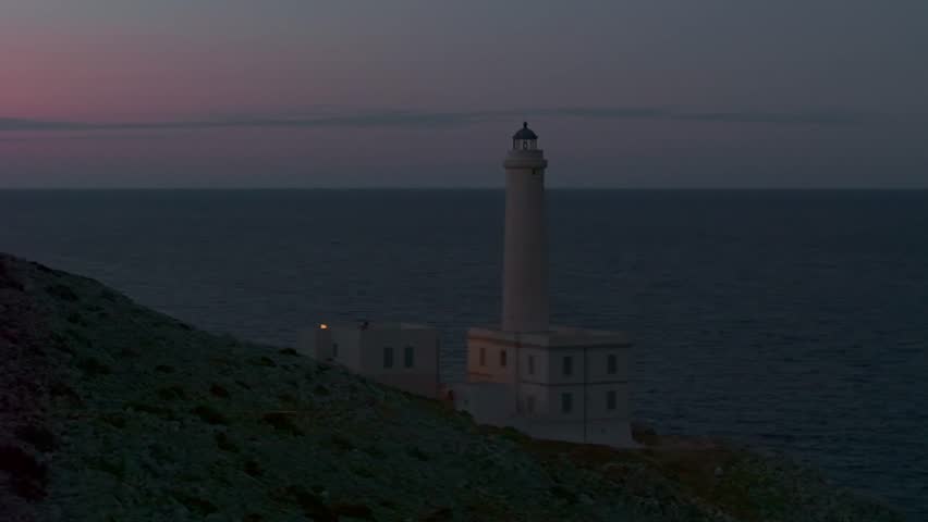 Aerial view of the historic Punta Palascia lighthouse at twilight. Features rotating light beams over the dark Adriatic Sea at Italy's easternmost point in the Salento region of Puglia.