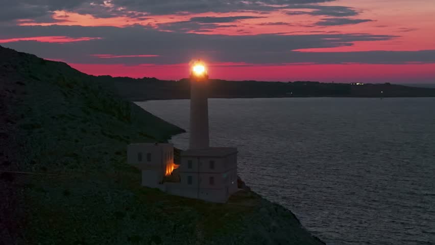 Aerial view of the historic Punta Palascia lighthouse at twilight. Features rotating light beams over the dark Adriatic Sea at Italy's easternmost point in the Salento region of Puglia.