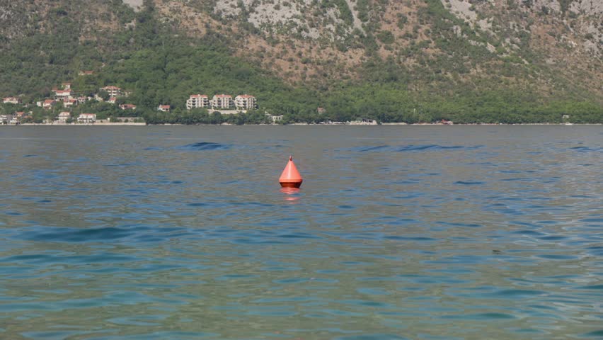 A bright orange conical buoy bobs on calm blue water as small waves shimmer. Red roof Mediterranean buildings sit below rocky tree covered slopes in midday light.