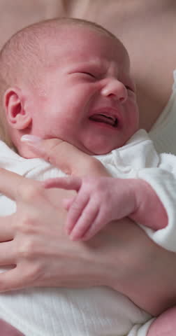 Mother cradles her distressed newborn baby, holding its tiny feet while it cries loudly