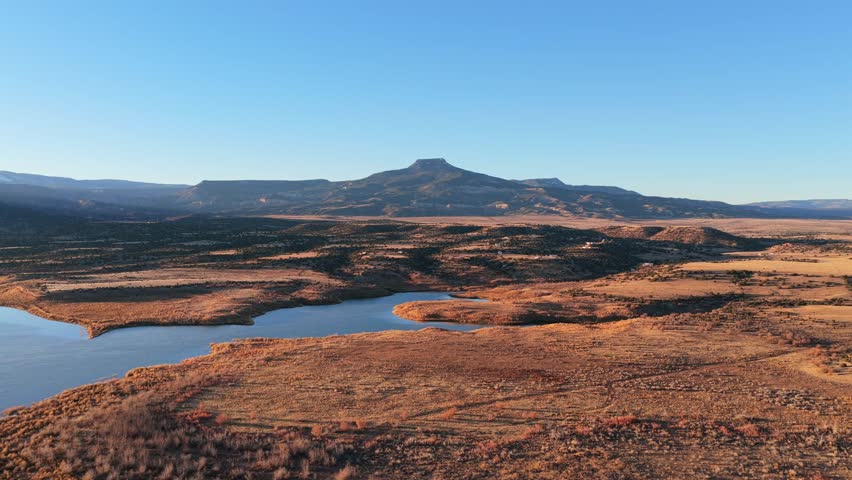 Aerial view captures the blue reservoir water of Abiquiu Lake below the volcanic basalt peak of Cerro Pedernal. Arid brush and sunlit shorelines define the high desert plateau of northern New Mexico.