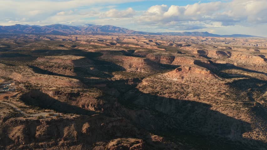 Patchy sunlight illuminates the tops of flat volcanic mesas while deep shadows fill the canyons below. This high desert landscape features arid vegetation and distant peaks under a cloudy sky.