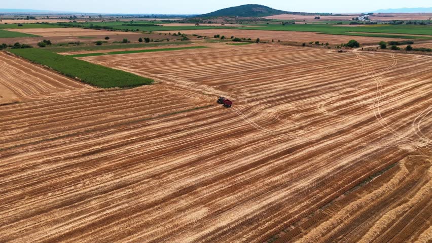 High-altitude aerial shot pushing and curving with a tractor and trailer moving through striped orange farmland, surrounded by endless agricultural fields and distant hazy hills.