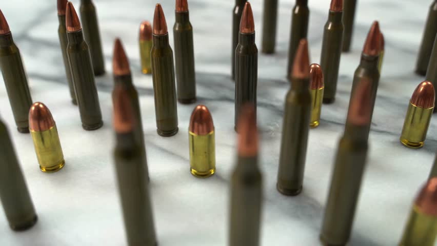 A macro slider shot moves across AR-15 components and rows of 9mm ammunition on a white table, highlighting polished metal, sharp geometry, and the mechanics of modern firearms.