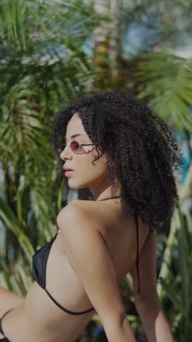 A stylish young woman with curly hair and sunglasses basks in the sun, surrounded by lush tropical foliage and a hint of blue water