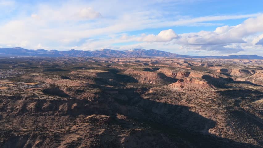 A wide aerial vista captures the expansive volcanic mesas and deep canyons of the Pajarito Plateau. Snow-capped mountain peaks sit on the horizon beneath a bright blue sky in northern New Mexico.