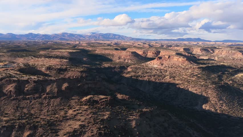 Wide aerial view captures expansive flat-topped mesas and deep canyon shadows in the New Mexico high desert. Snow-capped mountains sit on the horizon under a blue sky with white clouds.