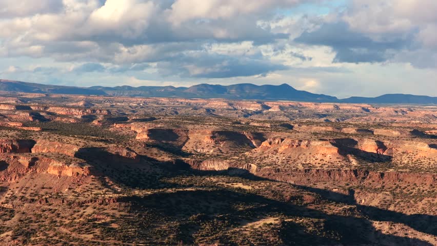 Dramatic afternoon light hits the flat volcanic mesas and deep canyons of the New Mexico high desert. Snow-capped mountains sit on the far horizon under a blue sky with white clouds in the winter.