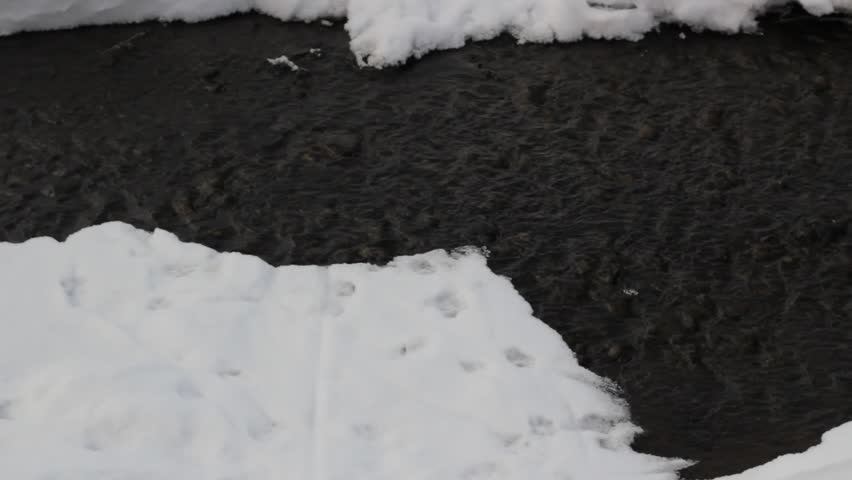 Static shot of a dark shallow stream flowing between white snowy banks. Detailed view of water ripples and pebbles in a peaceful winter nature setting.