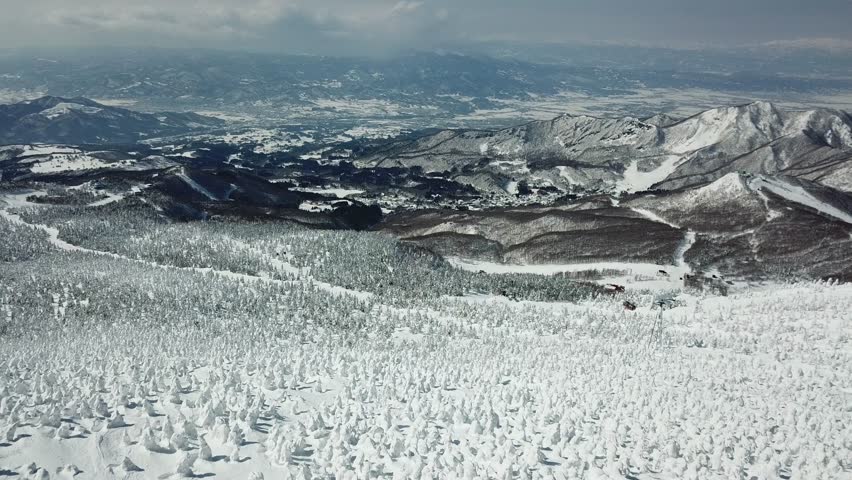 Aerial footage above snowy Mount Zao, where scenic cable cars fly over Juhyo forests (ice trees, snow monsters) on the slope, with a panoramic view of snow covered mountains toward the horizon on a cold winter day, in Zao hot spring and ski resort, Yamagata Prefecture, Tohoku Region, Japan.