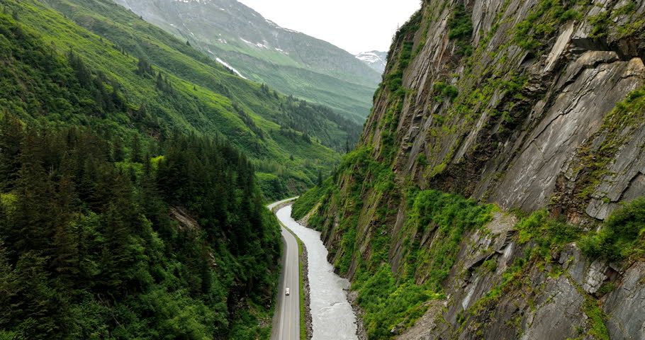 River and road at the foot of the unapproachable rocks. Journey by the beautiful wilderness of Alaska. Aerial view.
