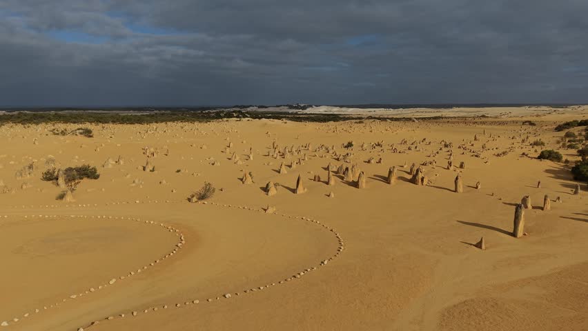 The surreal Nambung National Park Pinnacles Desert landscape featuring numerous limestone spires protruding from wind-swept sand. The scene captures rugged geology, vast openness, and the warm tones of late-day light contrasting with a moody sky.