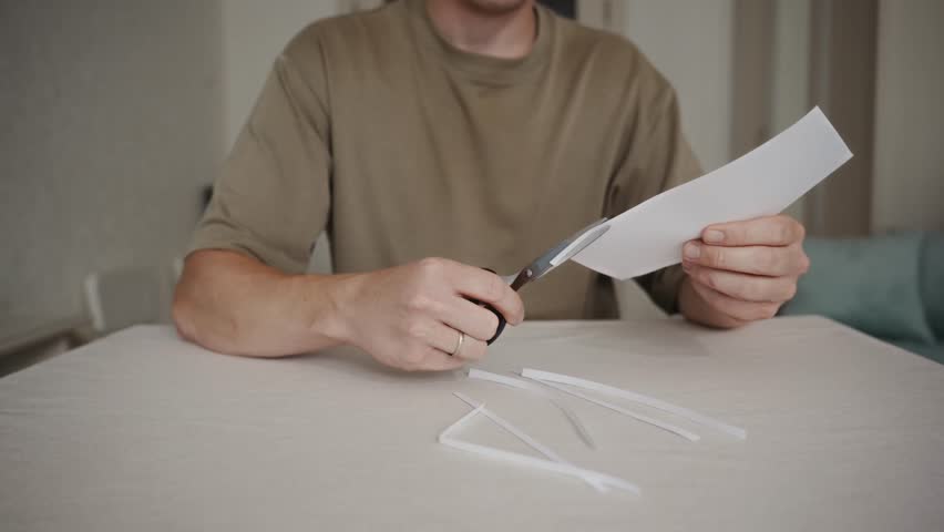 Man carefully cuts white paper with scissors while sitting at a table. Simple home environment and calm everyday action. High quality 4k footage
